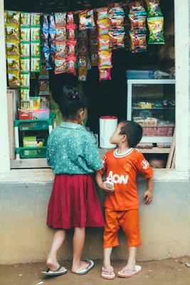 Two children stand in front of a small shop with various colorful snack packets hanging outside. The girl is wearing a green top and a red skirt, while the boy wears an orange shirt and shorts. The shop is filled with snacks and goods.