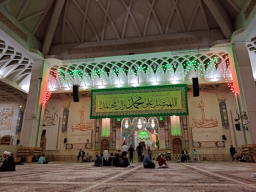 An ornate interior with a high ceiling and intricate architectural details, featuring Islamic calligraphy and decorations. The central area includes a large green banner with Arabic script, illuminated by green and red lights. People are sitting and standing on a patterned carpet, engaged in various activities.