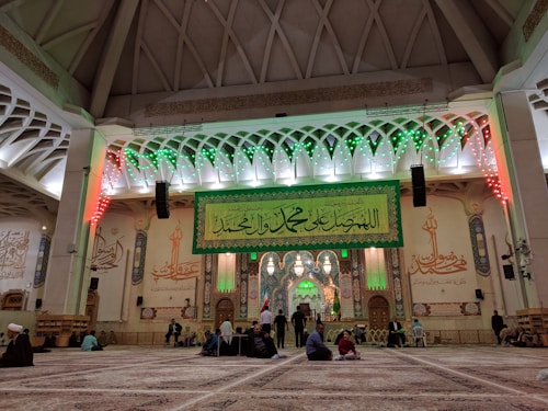 An ornate interior with a high ceiling and intricate architectural details, featuring Islamic calligraphy and decorations. The central area includes a large green banner with Arabic script, illuminated by green and red lights. People are sitting and standing on a patterned carpet, engaged in various activities.