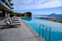 An infinity pool with clear blue water overlooks a scenic landscape, featuring mountains and a city in the distance. The poolside is lined with cushioned lounge chairs and neatly rolled towels. A modern building with glass balconies is adjacent to the pool area, and the sky is a bright blue with wispy clouds.