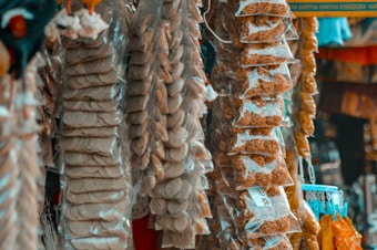 A market stall displays packaged snacks, with transparent bags containing various items like crackers and other snack foods hanging in rows. The colors are muted with a focus on earthy tones.