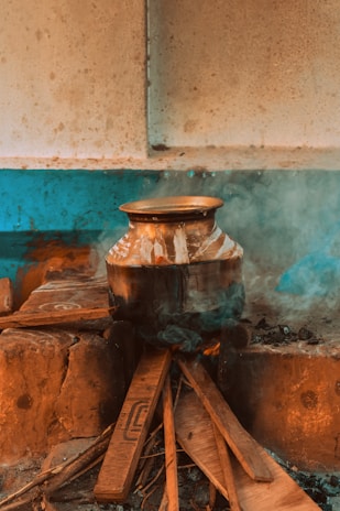 A metal pot is placed on a makeshift stove made of bricks and wooden planks. The stove is emitting smoke, indicating a fire is burning beneath. The setting appears rustic and traditional, with a blend of brick and wooden elements.