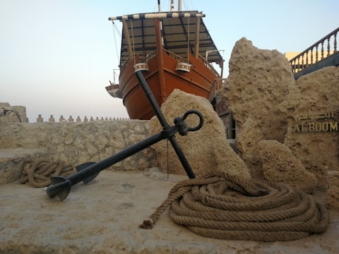 A traditional wooden ship with a large anchor in the foreground, surrounded by coiled rope and rocky structures. The ship is elevated in the background against a clear sky.