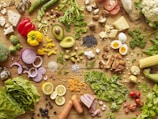 A variety of fresh vegetables, fruits, and other foods spread out on a wooden surface. Items include red and yellow peppers, sliced cucumbers, tomatoes, cauliflower, avocado, boiled eggs, various herbs, mushrooms, onions, broccoli, corn, lemon slices, rice, nuts, and cheese. The arrangement is colorful and organized to display the different textures and colors.