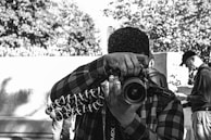 A person wearing a plaid shirt is holding a camera mounted on a flexible tripod, aiming the lens toward the viewer. The background includes other individuals who are blurred, focusing on different activities. Trees and a wall create an urban outdoor scene.