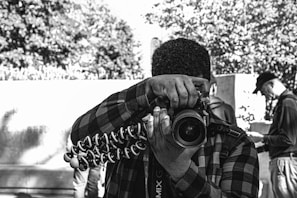 A person wearing a plaid shirt is holding a camera mounted on a flexible tripod, aiming the lens toward the viewer. The background includes other individuals who are blurred, focusing on different activities. Trees and a wall create an urban outdoor scene.