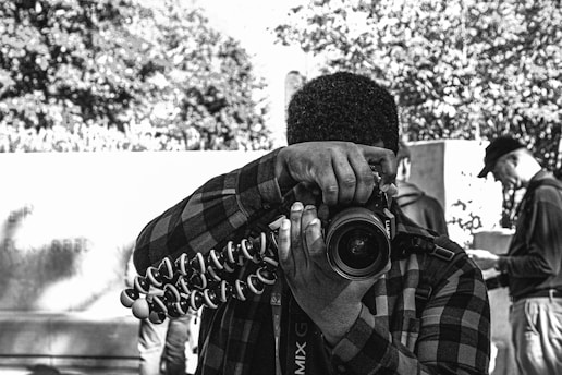 A person wearing a plaid shirt is holding a camera mounted on a flexible tripod, aiming the lens toward the viewer. The background includes other individuals who are blurred, focusing on different activities. Trees and a wall create an urban outdoor scene.
