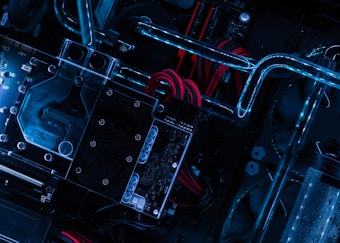 A close-up view of computer hardware featuring illuminated water cooling tubes winding over a motherboard, with visible circuits and red cables accentuating the setup.