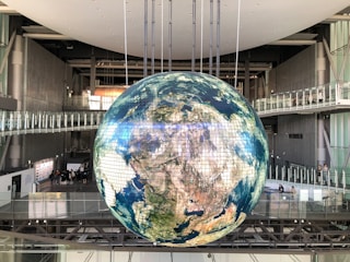 A large, spherical installation resembling a digital globe is suspended in a spacious, industrial-style indoor setting. The globe displays the Earth's continents and oceans, composed of pixelated panels. Metal structures, walkways, and glass railings surround the globe, with several people visible in the background.