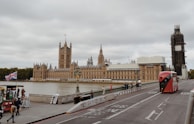 An iconic view of the Houses of Parliament in London, situated by the River Thames. The scene includes a red double-decker bus on the street, pedestrians walking on the sidewalk, and the British flag on display. The sky is overcast, adding a subdued lighting to the historic structure and its surroundings.