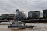 Several modern buildings line the riverside under a cloudy sky. A uniquely shaped, rounded building is central in the frame, with office buildings flanking it. In the river, two boats are visible, one is a white modern vessel marked with P272 and the other more elongated and white in color in the background.