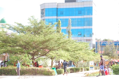 A bustling urban area featuring a modern building with a blue glass facade. In the foreground, there are lush green trees and shrubs. People are walking and interacting, some are seated in a shaded area under the trees. The presence of commercial signage in the background suggests a blend of natural and urban elements.