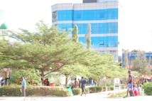 A bustling urban area featuring a modern building with a blue glass facade. In the foreground, there are lush green trees and shrubs. People are walking and interacting, some are seated in a shaded area under the trees. The presence of commercial signage in the background suggests a blend of natural and urban elements.