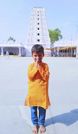 A young child stands barefoot on a paved area, wearing a bright orange traditional attire and blue pants. The child has hands clasped together as if in a greeting or prayer. In the background, a tall white temple with intricate architectural details is visible, along with some greenery and other smaller buildings.