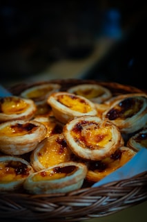 A woven basket filled with golden-brown pastries with glossy, caramelized tops, containing a rich custard filling. The pastries are stacked closely together, creating a warm, inviting look.