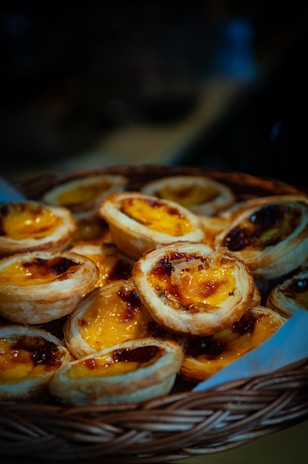 A woven basket filled with golden-brown pastries with glossy, caramelized tops, containing a rich custard filling. The pastries are stacked closely together, creating a warm, inviting look.