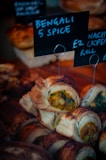 A close-up of a display of pastries filled with a colorful mixture of vegetables, labeled 'Bengali 5 Spice'. The pastries have a golden brown, flaky crust. Another sign next to it displays pricing information.