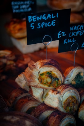 A close-up of a display of pastries filled with a colorful mixture of vegetables, labeled 'Bengali 5 Spice'. The pastries have a golden brown, flaky crust. Another sign next to it displays pricing information.