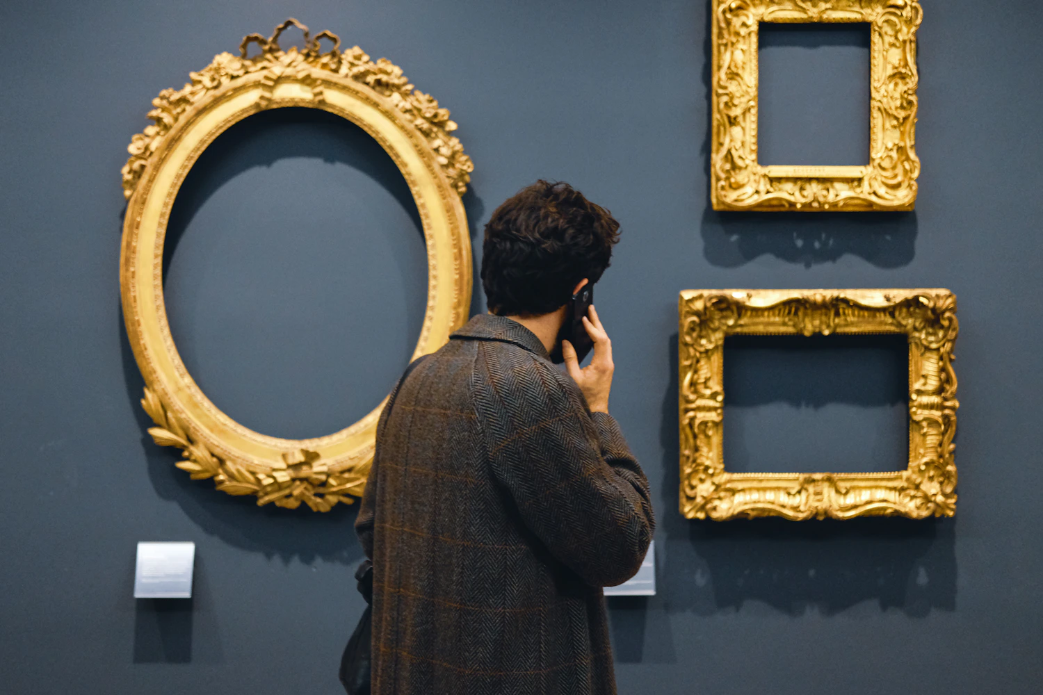 A man standing before three empty gilded frames on a dark grey wall