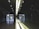 A spacious, modern interior with a reflective surface along one wall, creating a symmetrical appearance. The lighting is minimal, with ceiling lights and an illuminated strip along the wall. The floor is smooth and shiny, and the walls have a sleek, dark finish. Large glass doors at the far end allow some natural light to enter.