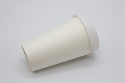 A plain, white disposable paper cup with a lid is positioned on its side against a neutral background. The cup appears to be clean and unused, suggesting it is for takeaway beverages.