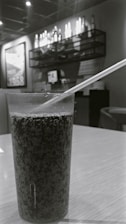 A glass filled with a dark, carbonated beverage is placed on a wooden table. In the background, there is shelving adorned with bottles and a framed poster. The setting appears to be an indoor cafe or restaurant with dim lighting.