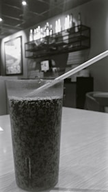 A glass filled with a dark, carbonated beverage is placed on a wooden table. In the background, there is shelving adorned with bottles and a framed poster. The setting appears to be an indoor cafe or restaurant with dim lighting.