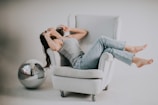 A person lounges comfortably on a light gray armchair, dressed in a gray tank top and blue jeans. They appear relaxed with their head tilted back, surrounded by a large, reflective disco ball resting near the armchair. The setting is minimalistic with soft lighting, creating a serene and casual atmosphere.