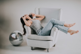A person lounges comfortably on a light gray armchair, dressed in a gray tank top and blue jeans. They appear relaxed with their head tilted back, surrounded by a large, reflective disco ball resting near the armchair. The setting is minimalistic with soft lighting, creating a serene and casual atmosphere.