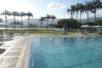 A large swimming pool with clear blue water is situated in an outdoor recreational area. Surrounding the pool, there are several lounge chairs, some shaded by umbrellas, with people relaxing on them. In the background, there are tall palm trees and mountains under a partly cloudy sky, which give the area a tropical and serene vibe. A lifeguard chair is positioned near the pool, and to the right, there are more seated individuals under larger canopies.
