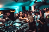 A lively nightclub scene with a crowd of people dancing and socializing. In the foreground, a DJ setup with turntables and mixing equipment is visible. The venue is dimly lit with blue and green lights, adding to the energetic atmosphere. People are engaged, with some looking directly at the camera, and others interacting with one another.