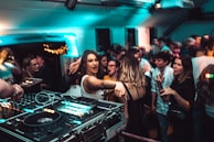 A lively nightclub scene with a crowd of people dancing and socializing. In the foreground, a DJ setup with turntables and mixing equipment is visible. The venue is dimly lit with blue and green lights, adding to the energetic atmosphere. People are engaged, with some looking directly at the camera, and others interacting with one another.