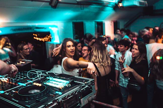 A lively nightclub scene with a crowd of people dancing and socializing. In the foreground, a DJ setup with turntables and mixing equipment is visible. The venue is dimly lit with blue and green lights, adding to the energetic atmosphere. People are engaged, with some looking directly at the camera, and others interacting with one another.