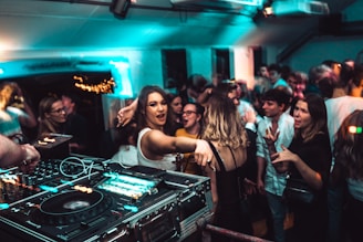 A lively nightclub scene with a crowd of people dancing and socializing. In the foreground, a DJ setup with turntables and mixing equipment is visible. The venue is dimly lit with blue and green lights, adding to the energetic atmosphere. People are engaged, with some looking directly at the camera, and others interacting with one another.