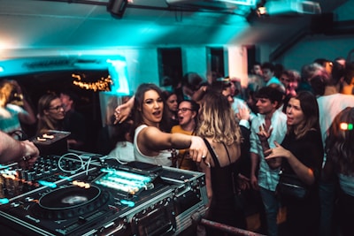 A lively nightclub scene with a crowd of people dancing and socializing. In the foreground, a DJ setup with turntables and mixing equipment is visible. The venue is dimly lit with blue and green lights, adding to the energetic atmosphere. People are engaged, with some looking directly at the camera, and others interacting with one another.