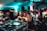 A lively nightclub scene with a crowd of people dancing and socializing. In the foreground, a DJ setup with turntables and mixing equipment is visible. The venue is dimly lit with blue and green lights, adding to the energetic atmosphere. People are engaged, with some looking directly at the camera, and others interacting with one another.