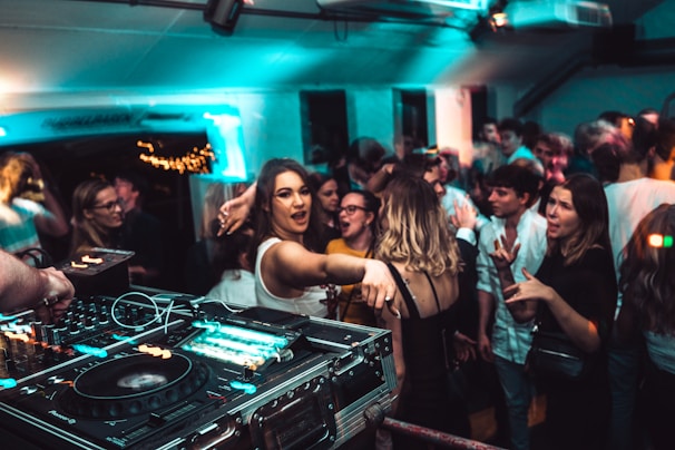 A lively nightclub scene with a crowd of people dancing and socializing. In the foreground, a DJ setup with turntables and mixing equipment is visible. The venue is dimly lit with blue and green lights, adding to the energetic atmosphere. People are engaged, with some looking directly at the camera, and others interacting with one another.
