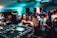 A lively nightclub scene with a crowd of people dancing and socializing. In the foreground, a DJ setup with turntables and mixing equipment is visible. The venue is dimly lit with blue and green lights, adding to the energetic atmosphere. People are engaged, with some looking directly at the camera, and others interacting with one another.