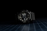 A dark-toned wristwatch with a robust and intricate design is centered against a black background. The watch face features multiple dials and markings, indicating complex functionality. The wristband is made of a durable material, and the overall appearance suggests ruggedness and reliability.