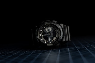 A dark-toned wristwatch with a robust and intricate design is centered against a black background. The watch face features multiple dials and markings, indicating complex functionality. The wristband is made of a durable material, and the overall appearance suggests ruggedness and reliability.