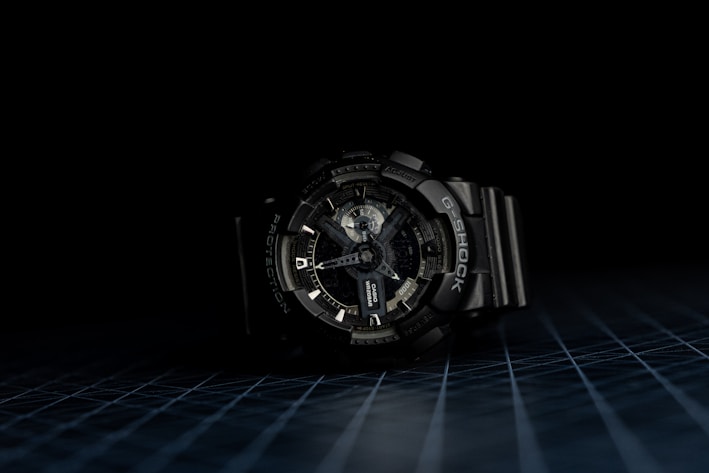 A dark-toned wristwatch with a robust and intricate design is centered against a black background. The watch face features multiple dials and markings, indicating complex functionality. The wristband is made of a durable material, and the overall appearance suggests ruggedness and reliability.