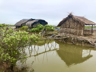 Two traditional huts with thatched roofs are situated near a body of water, surrounded by greenery. The huts appear rustic and are constructed with natural materials. A calm reflection of the structures and greenery is visible in the water.