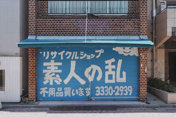 A storefront with a blue garage-style door featuring white Japanese text and a smiling cloud design. The building is made of brown bricks, and a small turquoise awning extends over the top of the door.