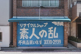 A storefront with a blue garage-style door featuring white Japanese text and a smiling cloud design. The building is made of brown bricks, and a small turquoise awning extends over the top of the door.