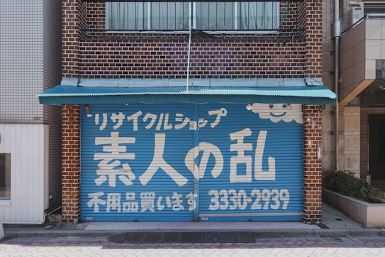 A storefront with a blue garage-style door featuring white Japanese text and a smiling cloud design. The building is made of brown bricks, and a small turquoise awning extends over the top of the door.