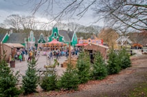 A lively outdoor scene features a colorful carousel surrounded by people in a festive market setting. Green gabled buildings create a quaint backdrop, with trees and neatly arranged spruce trees in the foreground. The atmosphere is bustling, with visitors exploring various stalls and attractions.