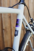 A close-up view of a bicycle frame showing the branding and design details. The frame is primarily a light gray color with a decal displaying the words 'Stingray' and 'R2'. The text is accented with purple and black elements. The bicycle appears to be leaning against a wooden background.