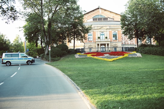 A historic building with a symmetrical facade is situated behind a well-maintained garden featuring a floral design with red, yellow, and white flowers. The building is partially obscured by trees, and there is a paved road leading up to it. A police van is parked on the road, adding an element of security or authority to the scene.