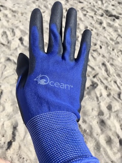 A blue and black glove with a text logo on it, worn on a hand, is shown against a sandy beach background. The glove is textured for grip, and the beach surface has visible footprints and small ridges.