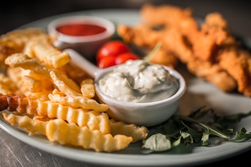 A plate featuring crinkle-cut fries, breaded and fried chicken strips, a small bowl of creamy sauce, and fresh cherry tomatoes. Garnishes include greens such as arugula, adding a touch of color and freshness.